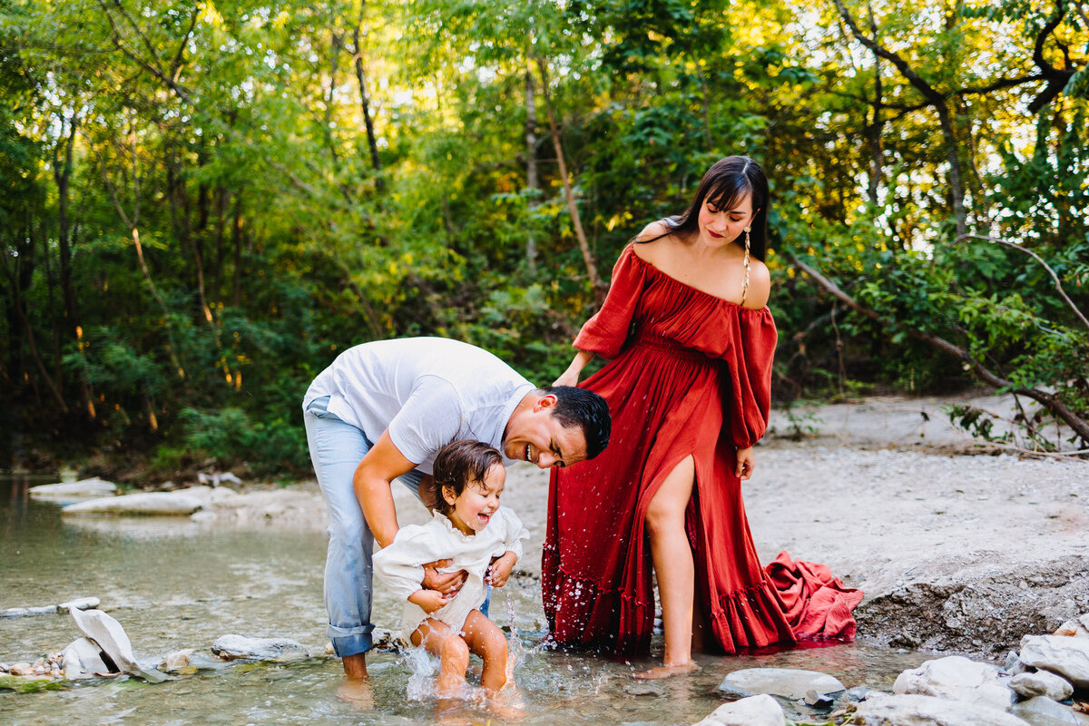 A beautiful family is taking a mini session  photography on the river. The mom is wearing a long red dress and the baby boy is playing with the water