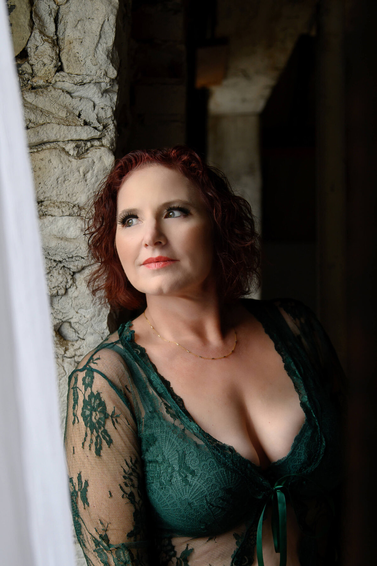 Red haired woman in green bra leaning against a brick wall looking out the window for her Hamilton Boudoir Photography session