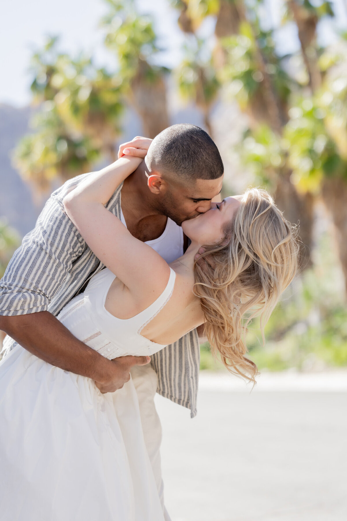  Romantic image of a couple mid-dip and kiss, surrounded by soft sunlight and greenery in a joyful engagement-style pose.