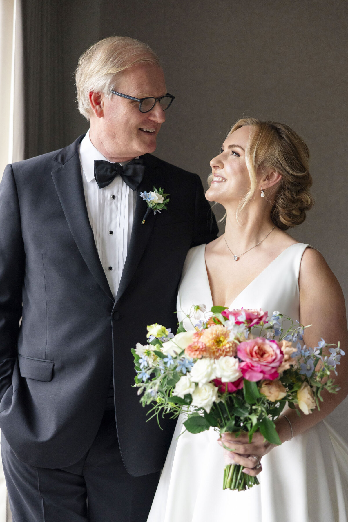 A bride in a white dress holds a colorful bouquet and looks at a man in a black tuxedo. They are standing indoors, smiling at each other, in front of a window with soft lighting.