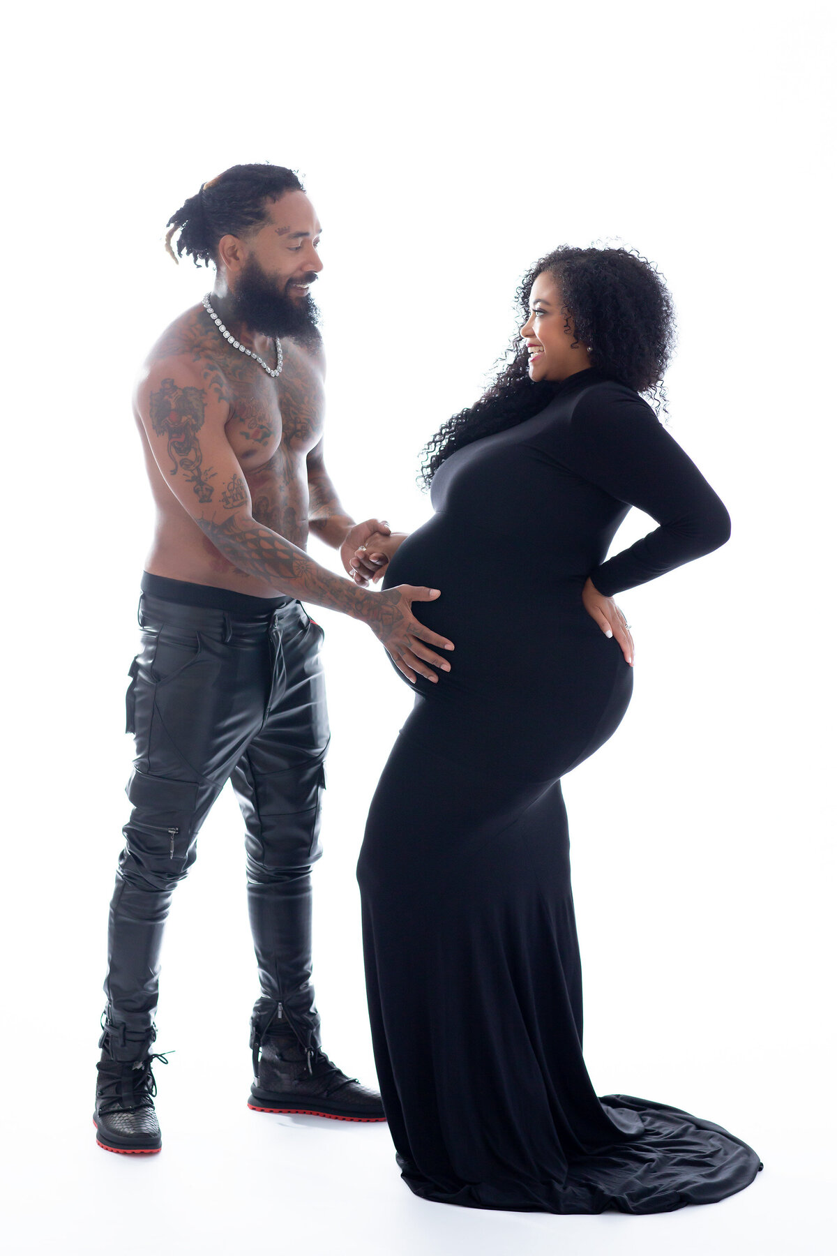 A smiling couple. Dad is shirtless wearing fashionable black leather pants with tattoos on his chest and arms. HIs happy wife is wearing a fitted black wife and his hands rest on her belly. Maternity photography in Dallas by Lindsay Walden.