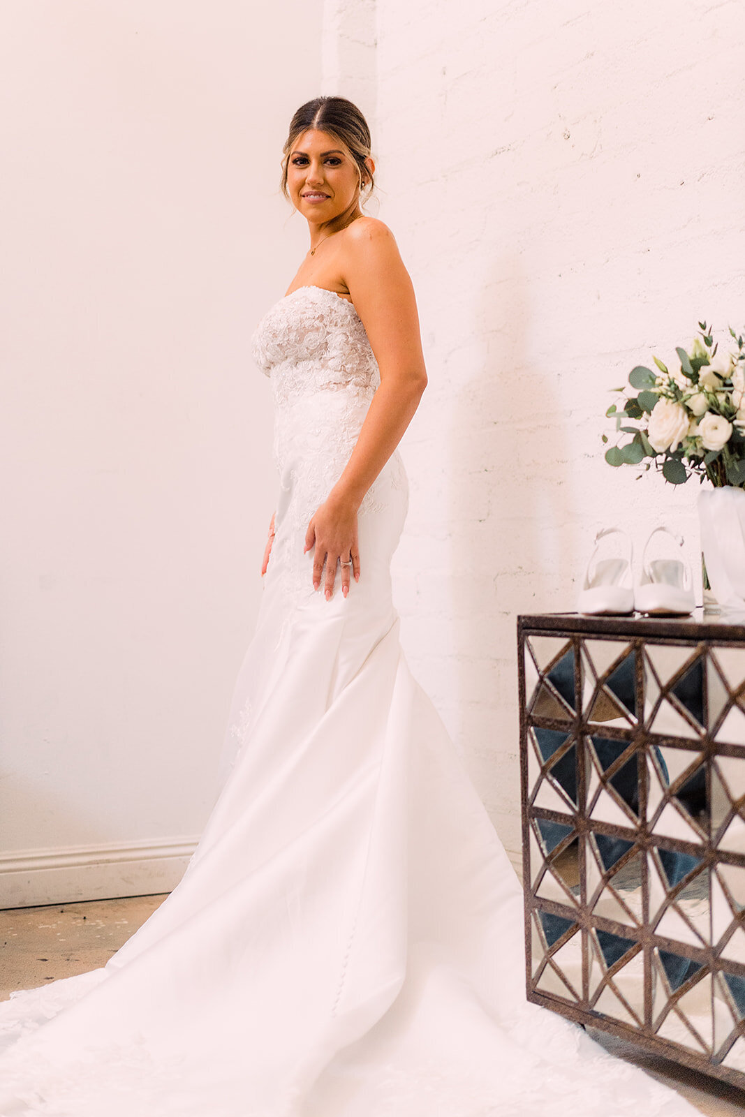 The bride poses in her strapless wedding gown with floral lace details, standing next to a mirrored cabinet with a bouquet and shoes displayed.