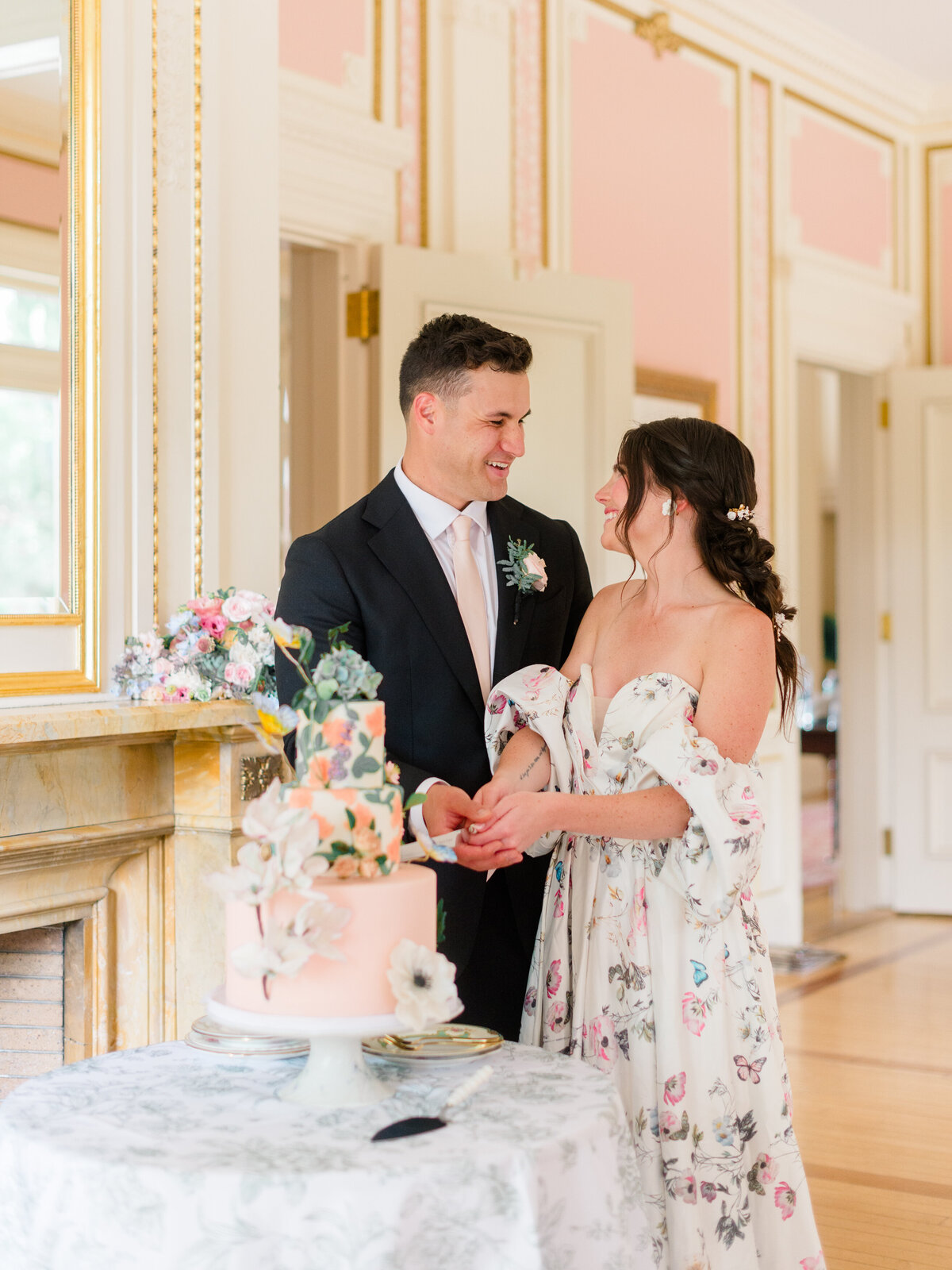 Bride and groom share a joyful moment cutting a hand-painted floral wedding cake at Cairnwood Estate, surrounded by romantic pastel decor and French-inspired elegance. Captured by Elizabeth Kane Photography, Philadelphia Fine Art Wedding Photographer, this image blends luxury wedding storytelling with timeless, emotive detail.