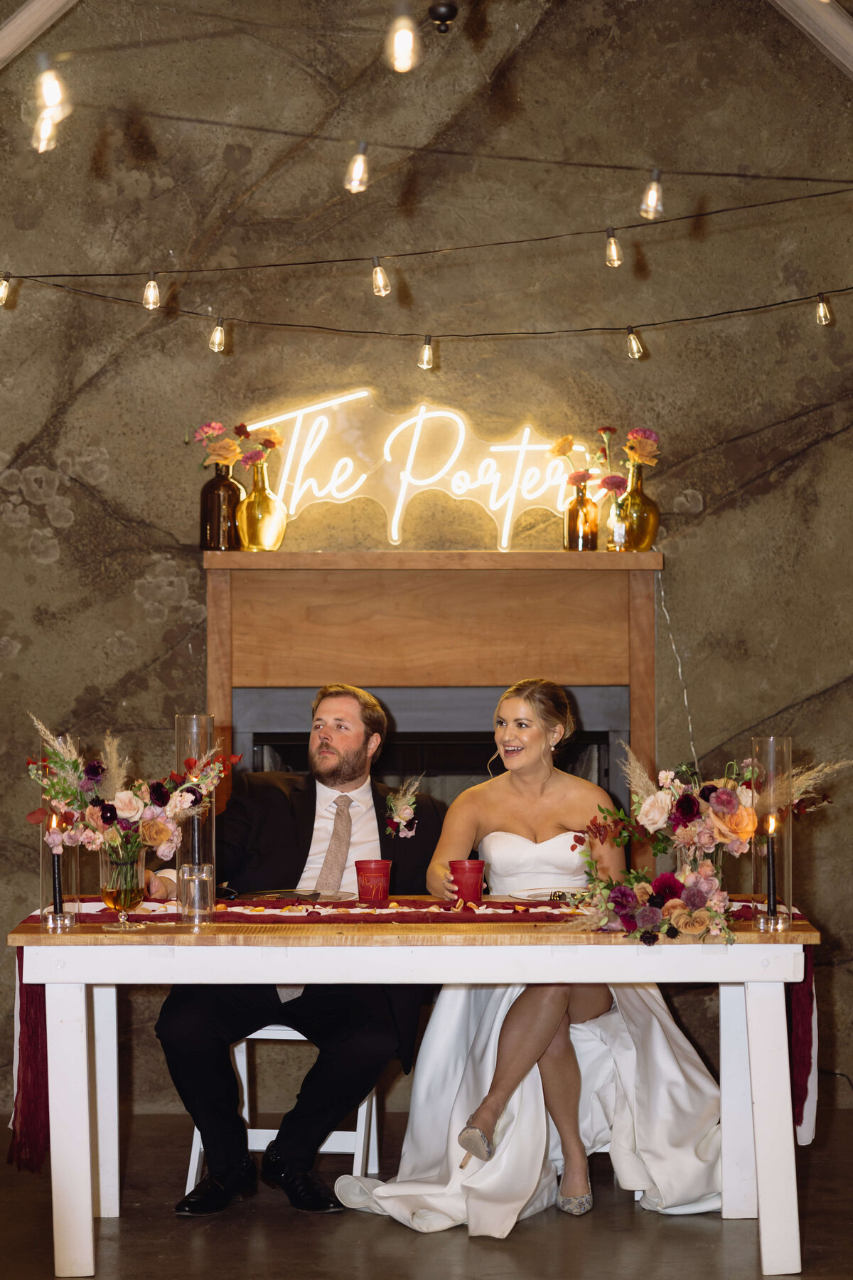 wedding reception with bride adn groom sitting at their wedding table that is decorated with florals and red table runner with their last name in a light sign above a mantle behind them