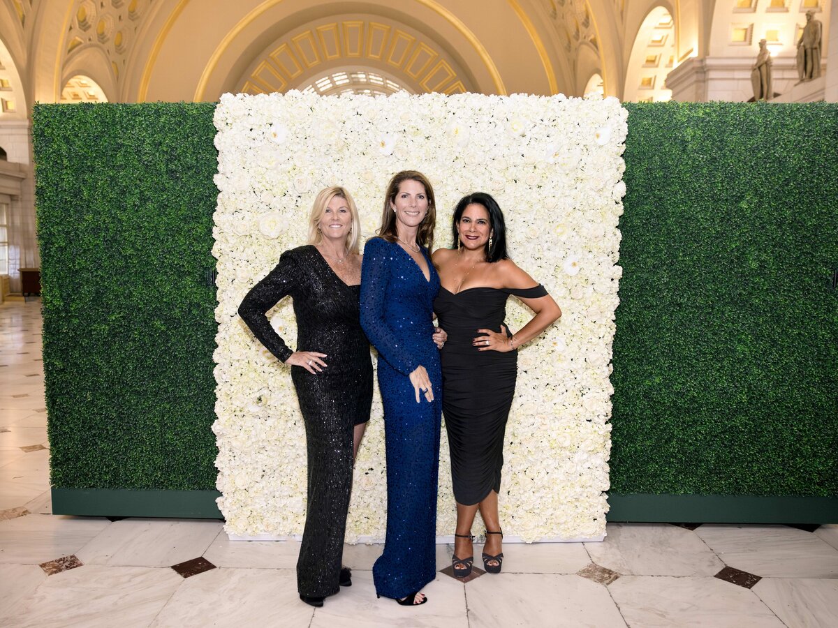 Three women in elegant dresses stand in front of a backdrop made of white flowers and green foliage. The setting is indoors, with an arched ceiling and ornate architecture visible in the background.