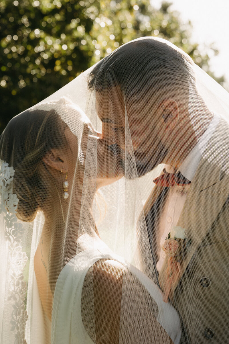 Mariés s'embrassant sous un voile dans un décor naturel. Mariage imaginé par Nos Moments Ensemble, wedding planner Nantes Loire Atlantique Vendée.