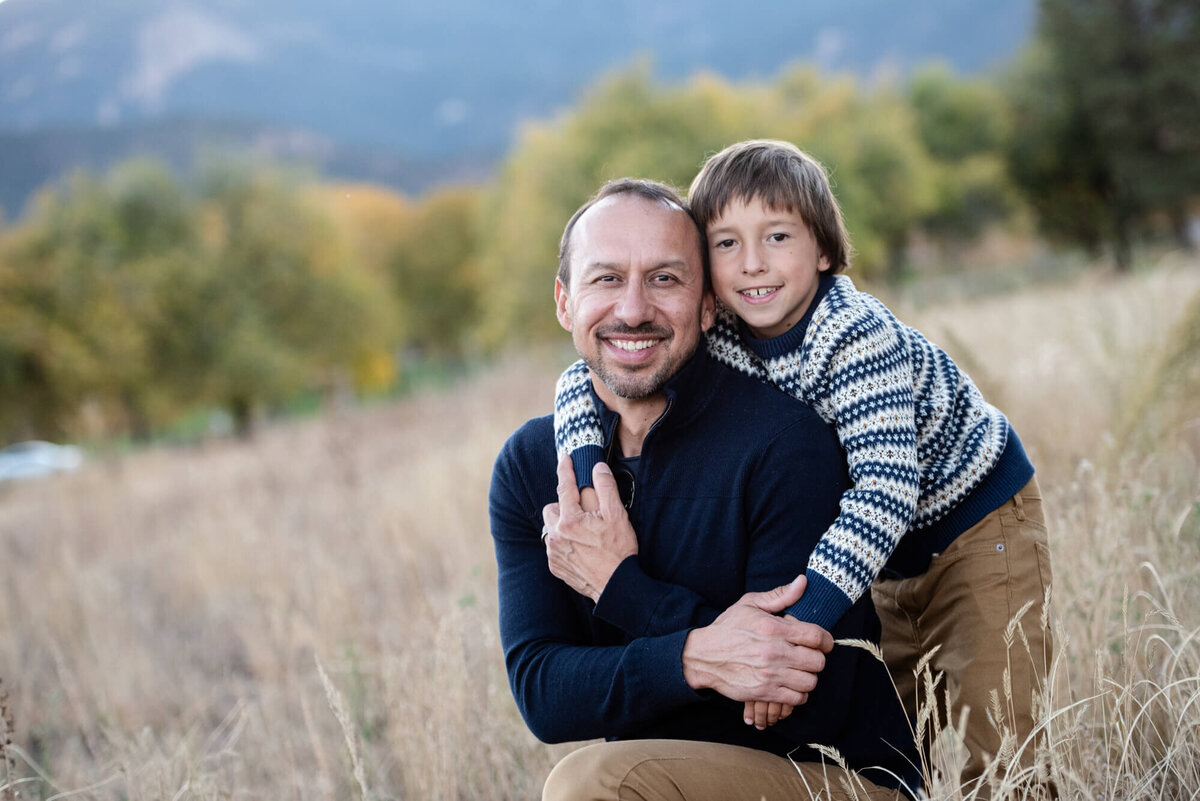 A boy in a stripe sweater hugs his dad while in a park field of golden grass