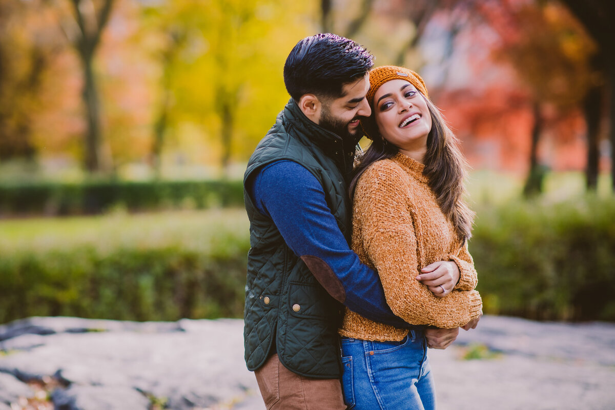 An engaged couple embrace wearing autumn clothing at central park in the fall in nyc