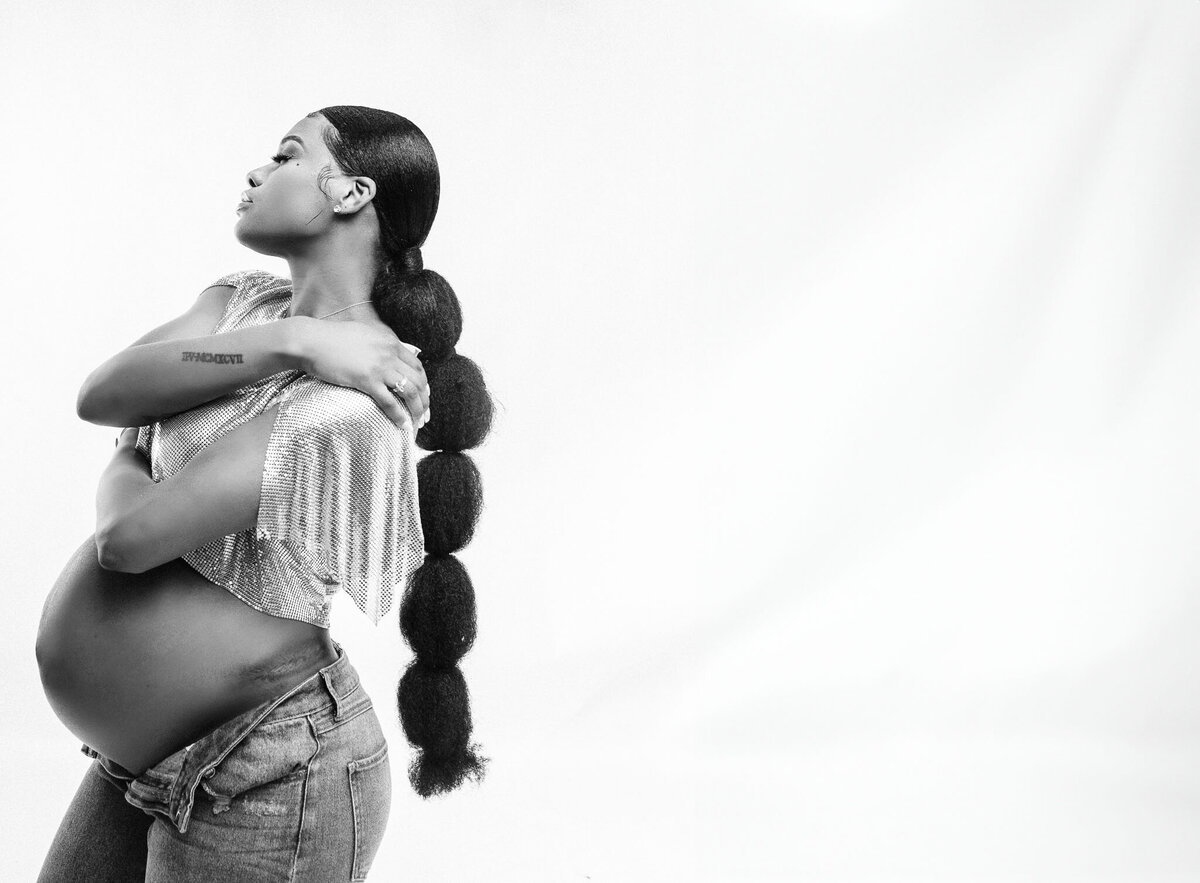 A striking black and white maternity photo of a Black mother in Columbia, SC, wearing a long ponytail, jeans, and a chic chainmail top. A bold and artistic representation of motherhood.