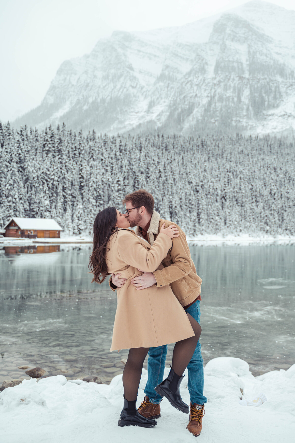 Snowy kiss in front of log cabin and icy lake in Lake Louise engagement session.