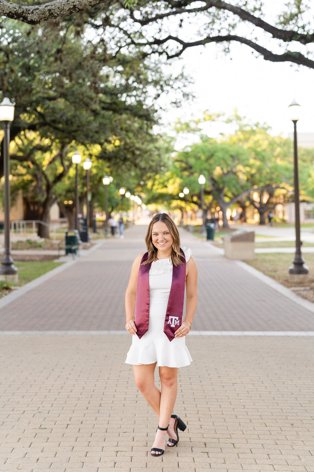Texas A&M senior girl holding Aggie stole and wearing white dress in the trees of Military Plaza