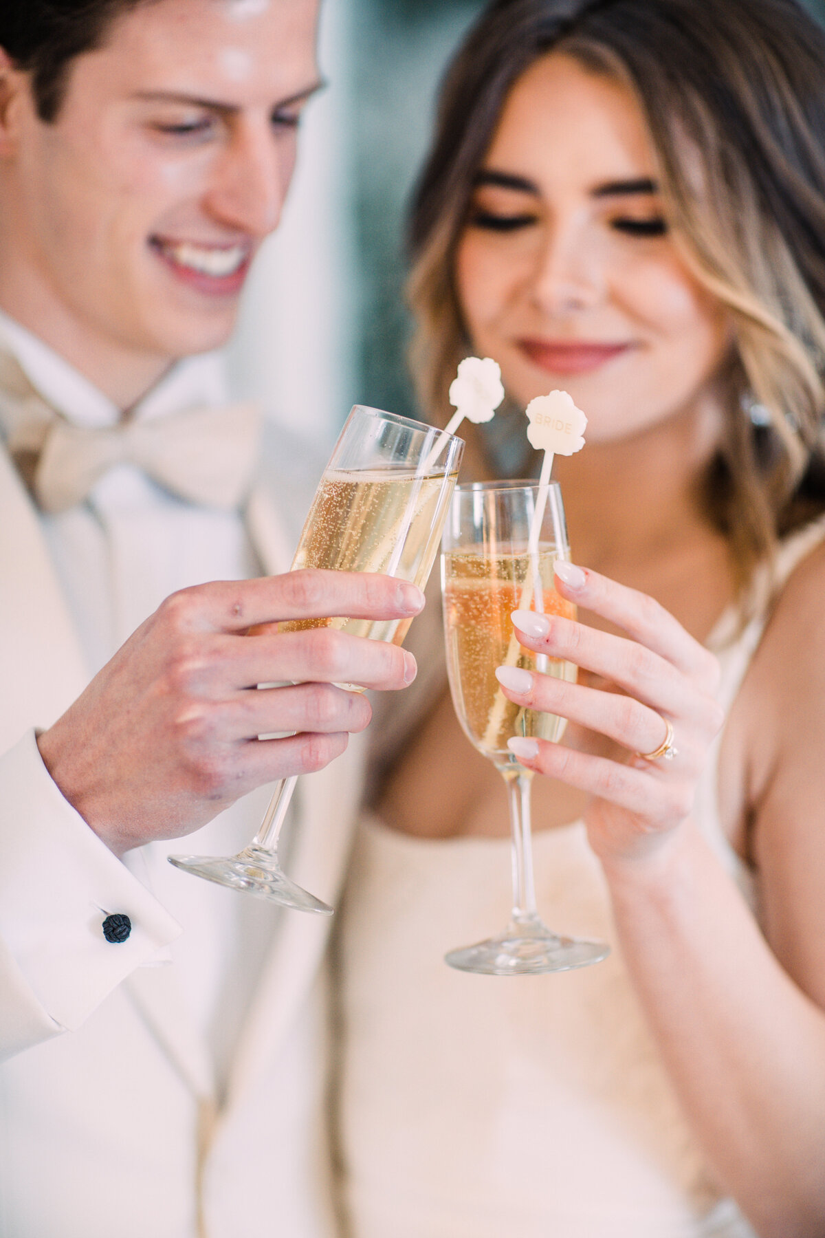Groom in white tuxedo & bow tie  & bride toast with glasses of champagne with acrylic drink stirrers