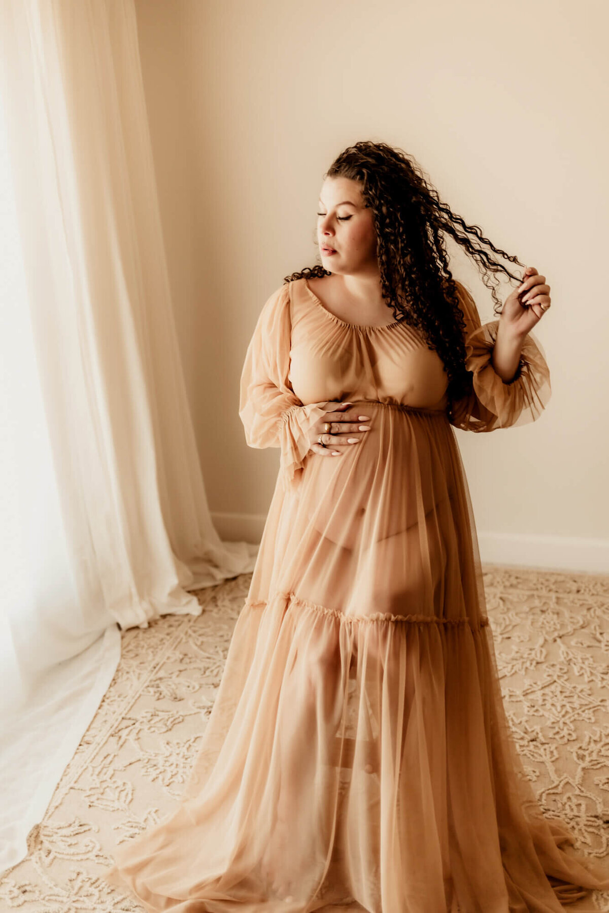 Woman with curly hair plays with it while placing her hand on her baby bump and sitting on a stool, Norman Oklahoma.