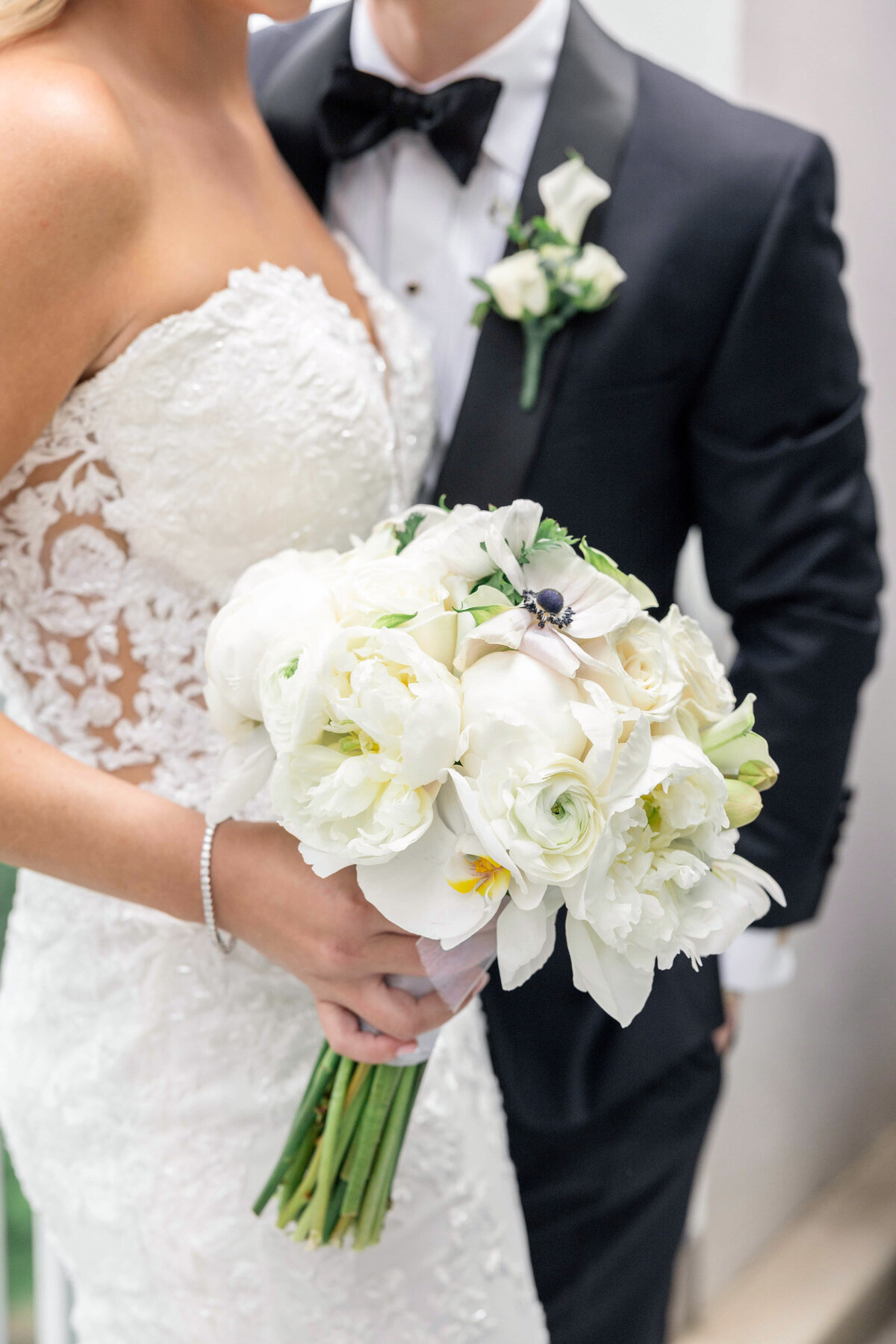 A bride and groom in elegant wedding attire stand closely together. The bride holds a bouquet of white flowers, wearing a strapless lace gown. The groom is in a black tuxedo with a white boutonniere.