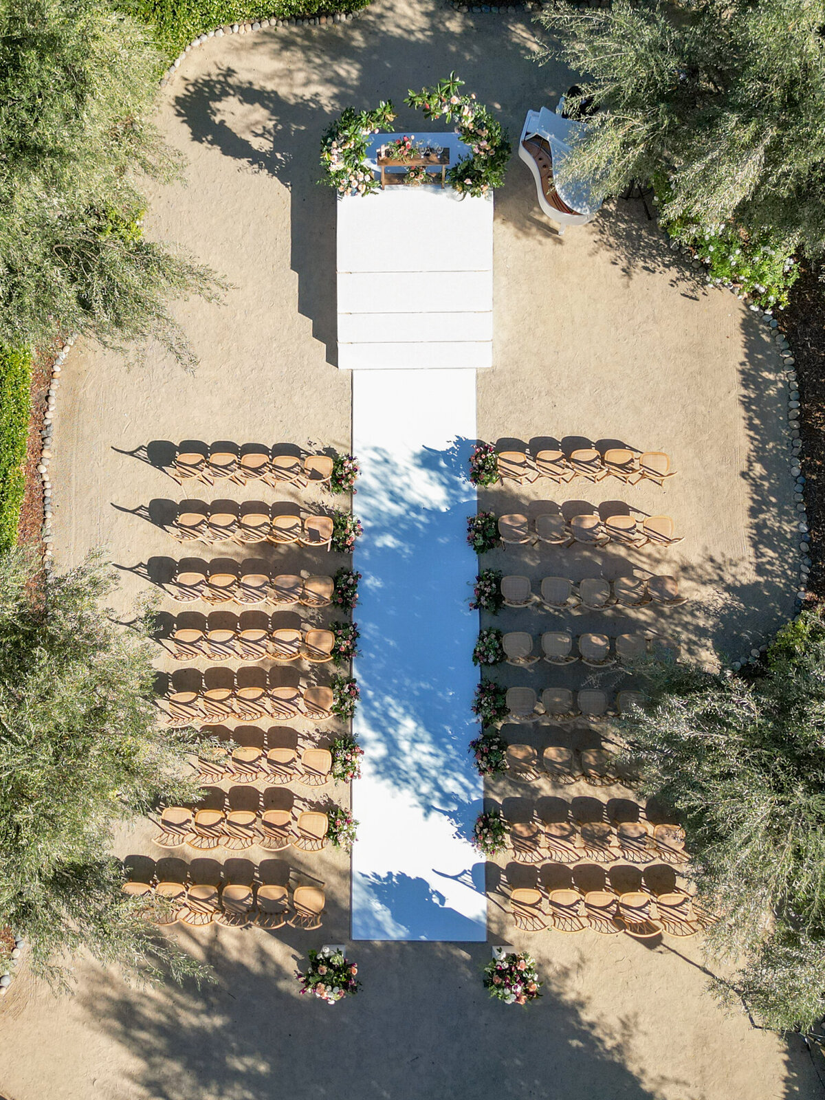 Aerial view of an elegant outdoor ceremony setup—neatly arranged chairs, floral-lined aisle, and sun-drenched vineyard charm.