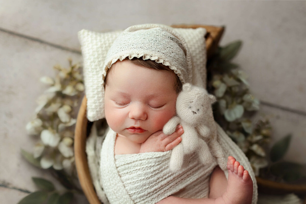baby newborn wearing a white wrap and sleepy cap holding a knit bear laying in a wooden moon shaped bowl
