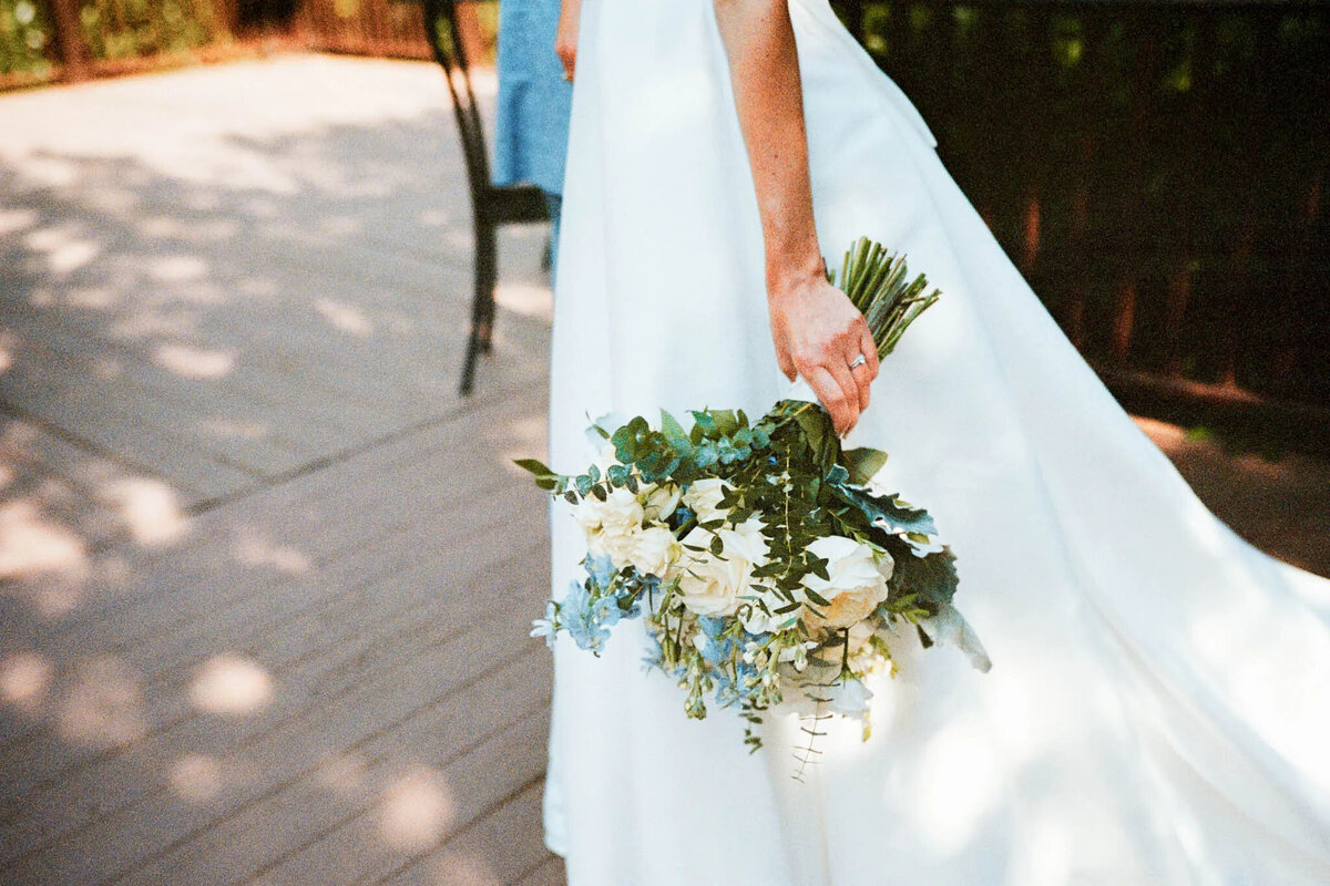 A bride in a white dress holds a bouquet of white and light blue flowers with greenery, standing outdoors on a wooden deck—captured beautifully by an NJ wedding photographer. Only her lower torso, arm, and bouquet are visible.