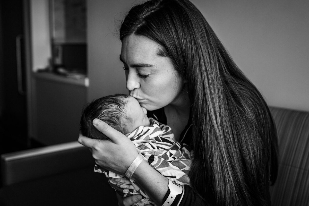 mom holding swaddled newborn close and kissing his forehead