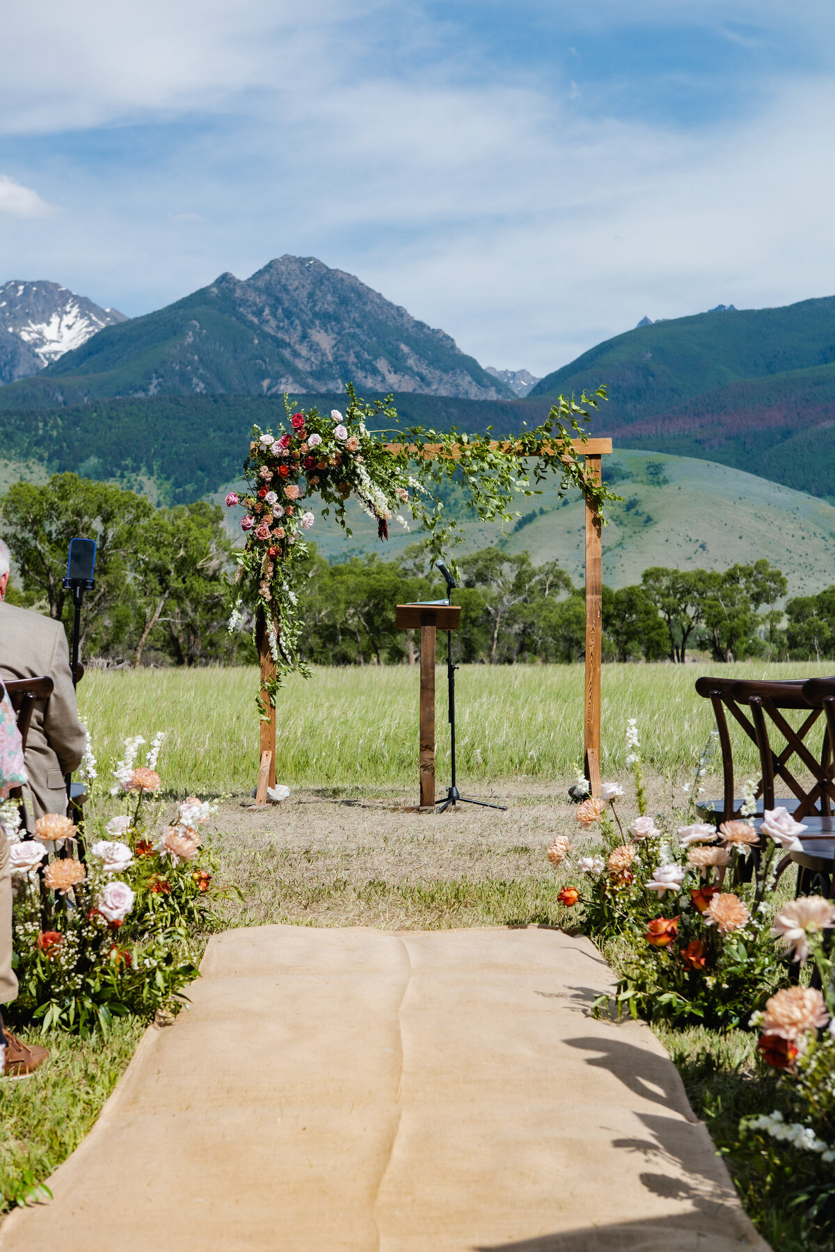 rustic mountain wedding ceremony setup featuring a wooden arch adorned with garden roses and cascading greenery, floral aisle arrangements, and scenic Montana peaks in the background