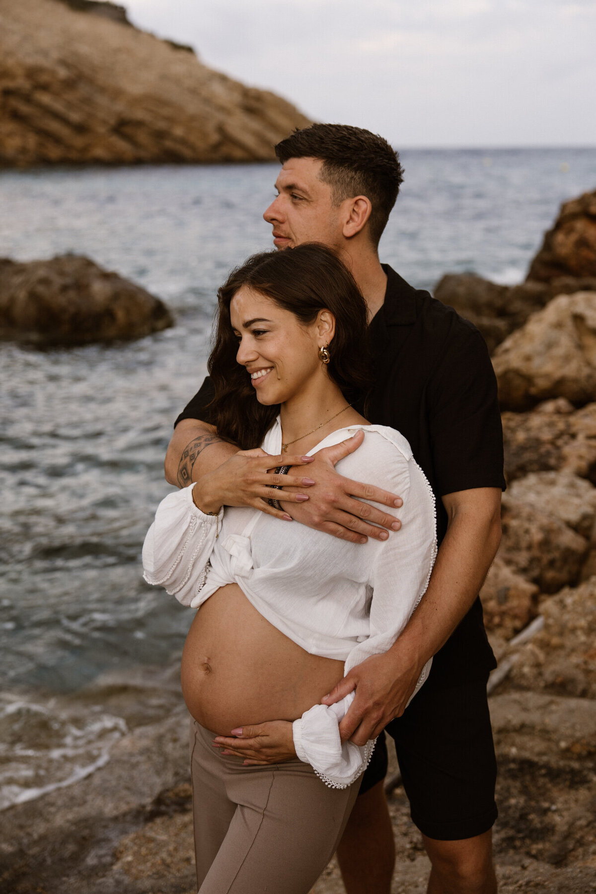 Man is standing behind a woman and holding her while she poses with belly during pregnancy photoshoot on Ibiza