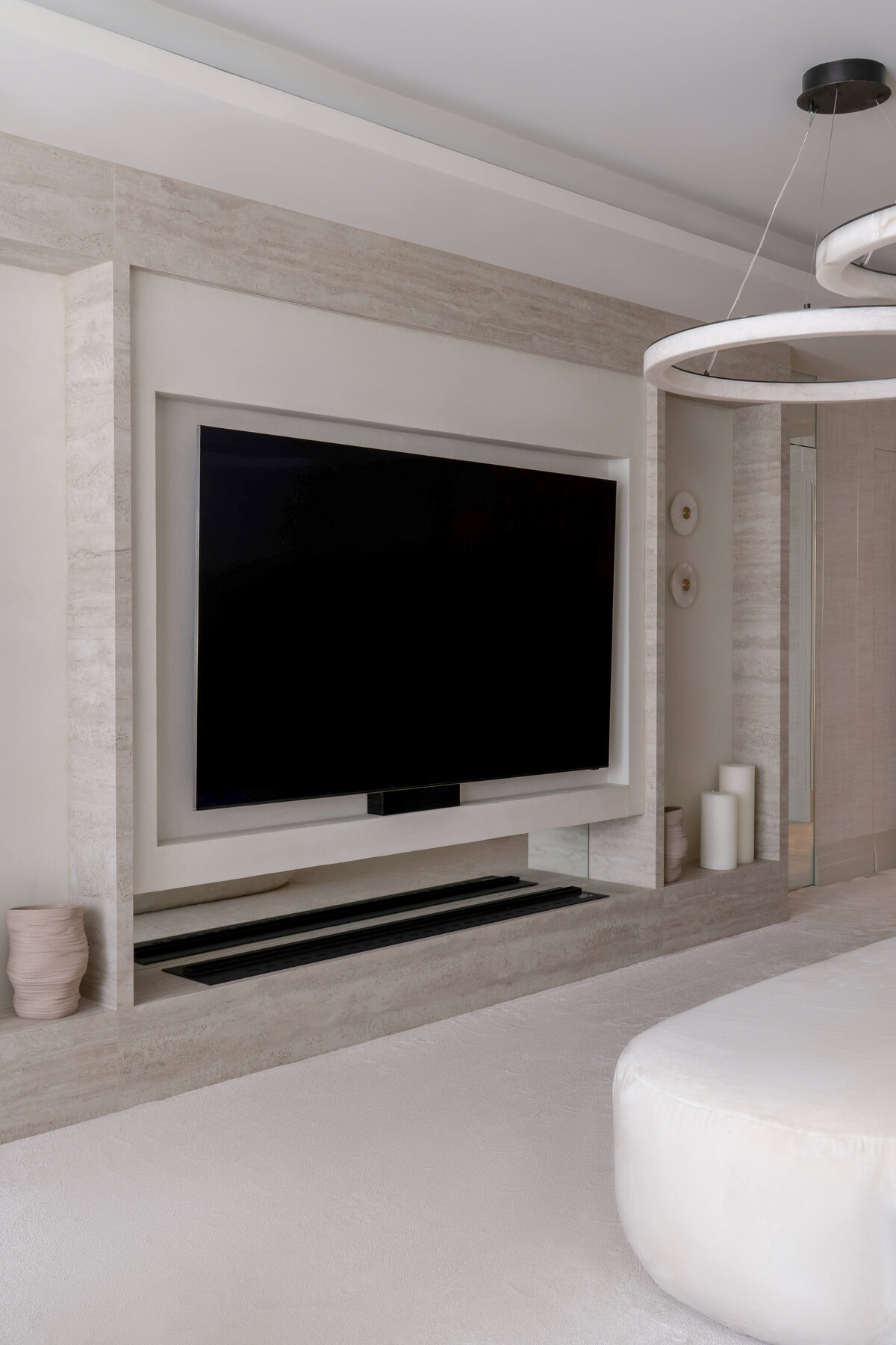 A large television recessed in the wall opposite the bed in a large primary bedroom with a chic, neutral aesthetic.