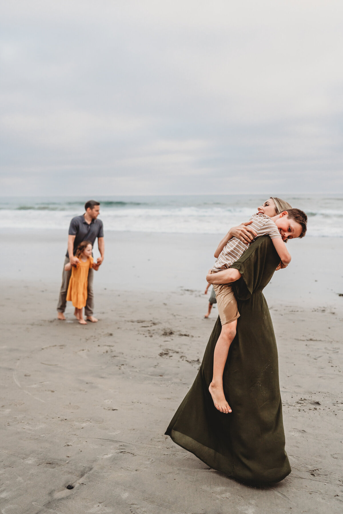 Mother holds and embraces her son in her arms with ocean waves in the background at Del Mar beach, San Diego family photography.