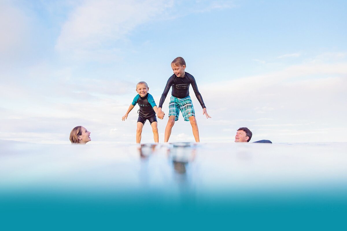 Two boys photographed by Love + Water stand on surfboards in the ocean on Maui as parents laugh and look up at them