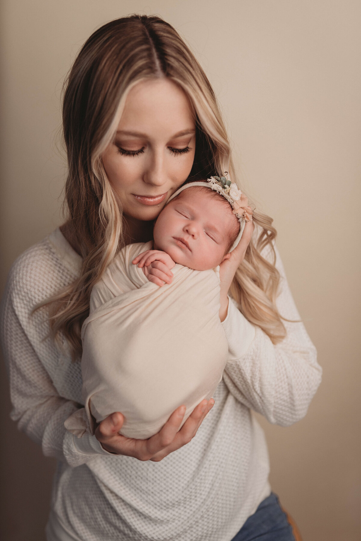 Blonde hair mom wearing white standing holding newborn baby girl close to her cheek snuggling her with closed eyes.