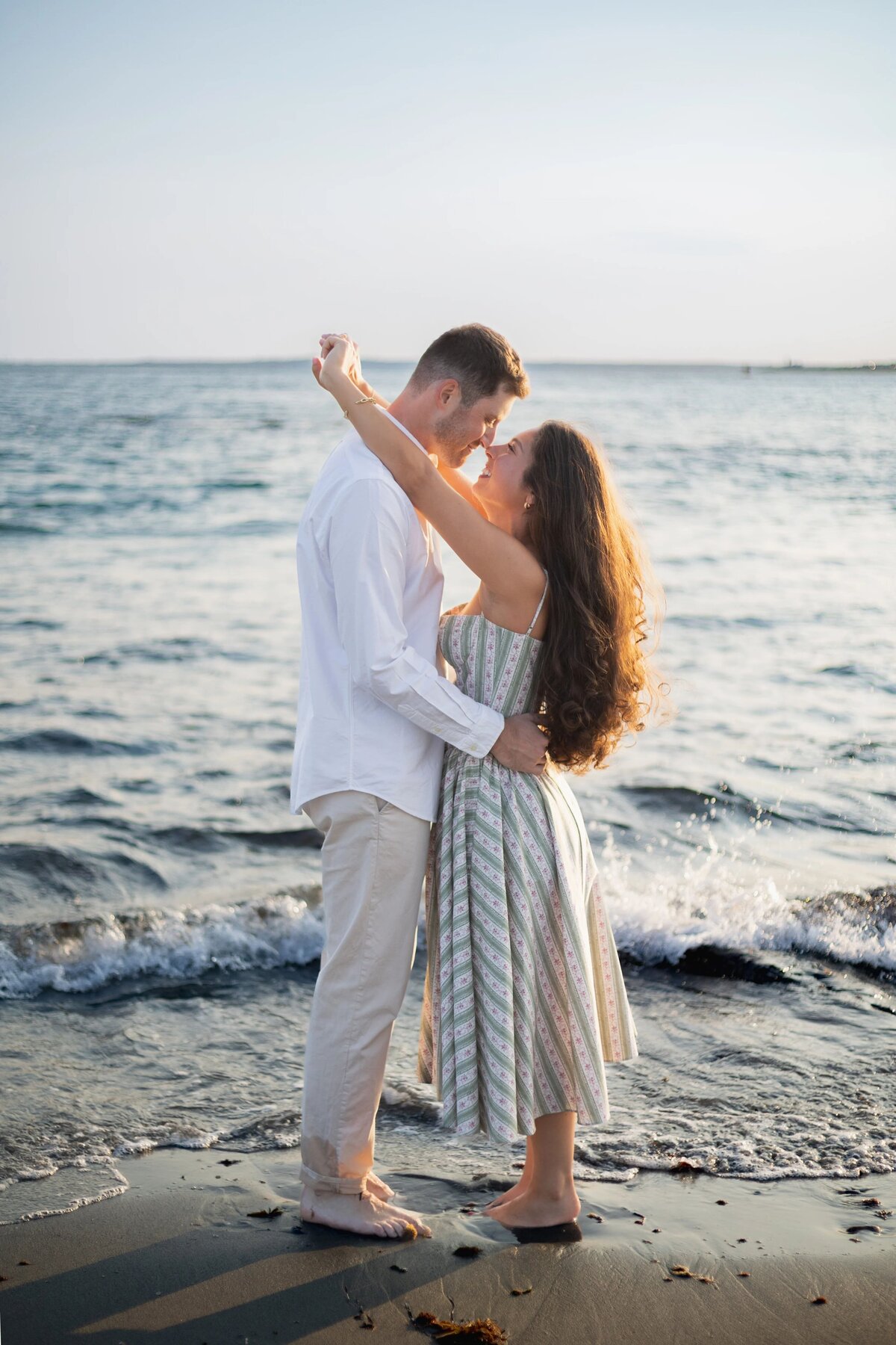 Wedding Photographers in RI | Kelsey Sheehan Photography Timeless Rhode Island Weddings | A couple embraces lovingly by the ocean, with gentle waves in the background. The woman wears a striped dress; the man is in a white shirt and beige pants.