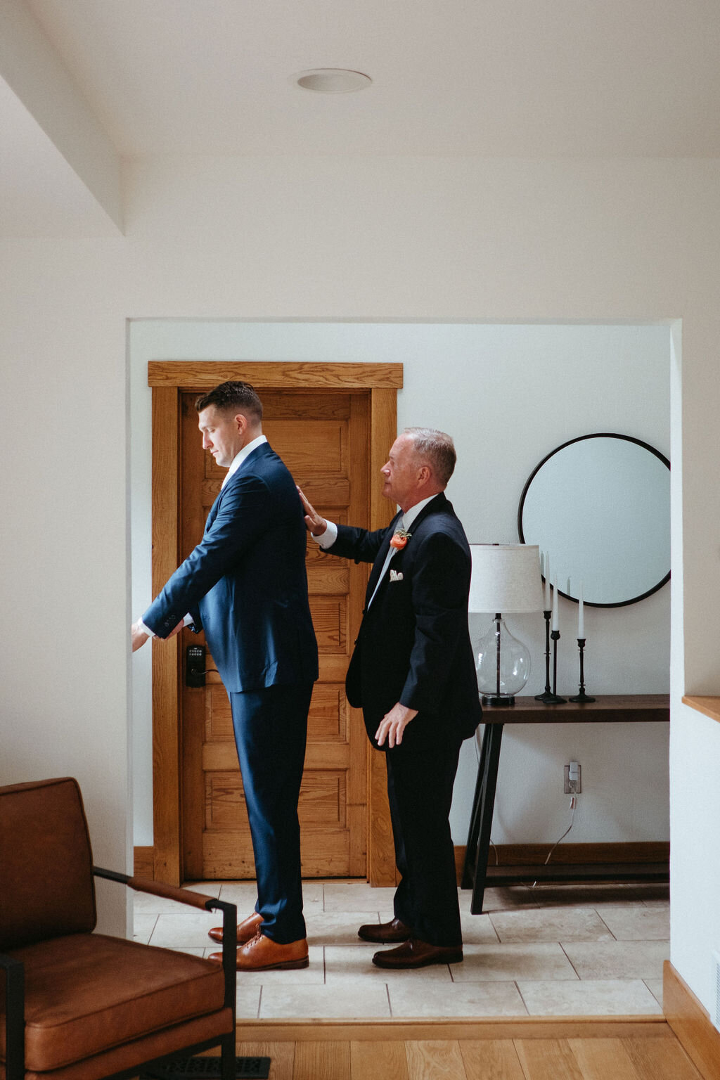 the father of the groom assisting his son in putting his suit jacket on for his wedding at Willowbrook wedding venue in the modern farmhouse entry hallway