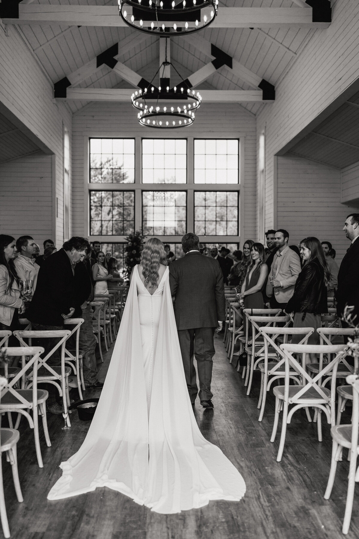 A black and white photograph of Carly and her father on their wedding day at Morgan Creek Barn in Dripping Springs, TX. The viewpoint is from behind the bride and her father as they walk down the aisle toward the ceremony. Her white wedding dress has wings from each shoulder that flow to the floor and trail behind her. Wedding photography by Stacie McChesney of Vitae Weddings.