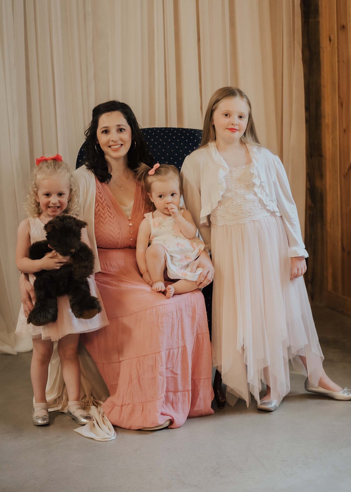 Maddie Rae Photography mom and her three daughters. Mom is sitting in the chair with baby girl in her lap, the other two girls are standing next to her, one is hugging a teddy bear