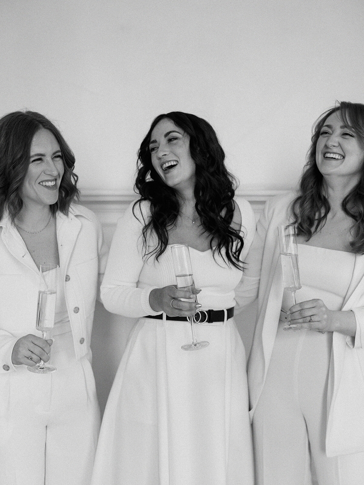 Three women, dressed in white, smile and laugh while holding glasses of sparkling wine, celebrating a memorable event curated by Destination Wedding Planner Melissa Dawn Event Designs.