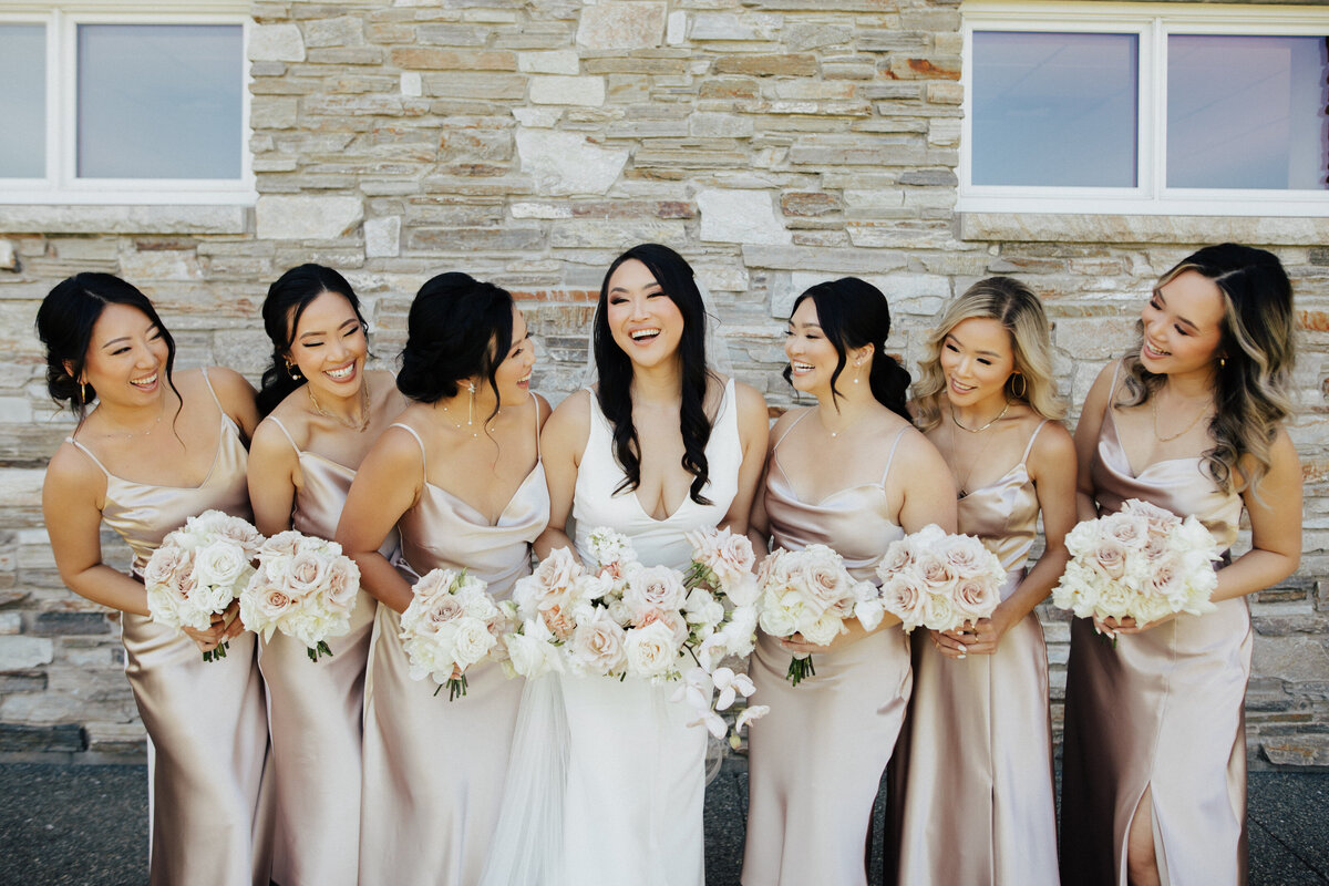 Stunning bride laughs with her bridesmaids on her wedding day while holding floral bouquets