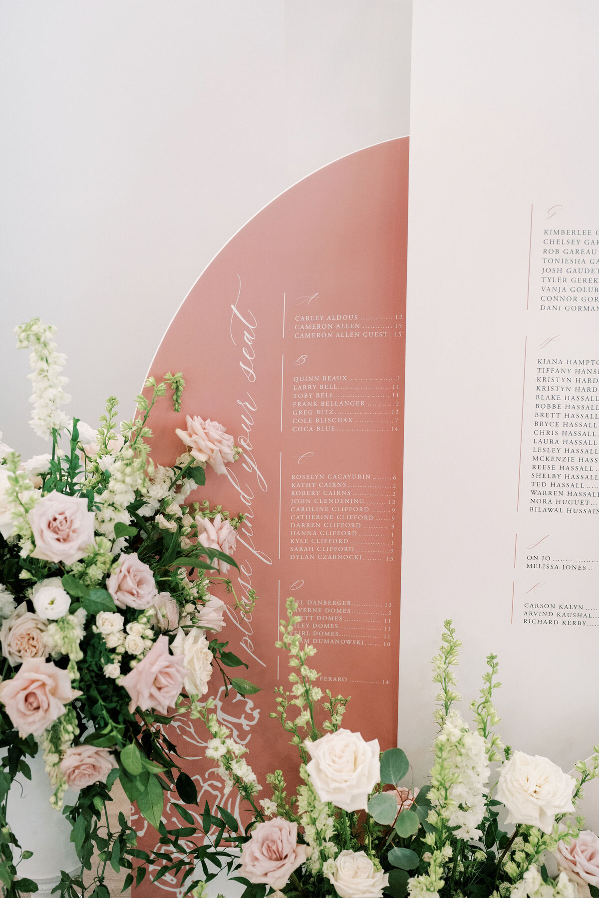 A wedding seating chart on a rose-colored background with names and table assignments in white text, adorned with white and light pink flowers, perfectly captures the essence of a romantic Norland Estate wedding in Calgary.