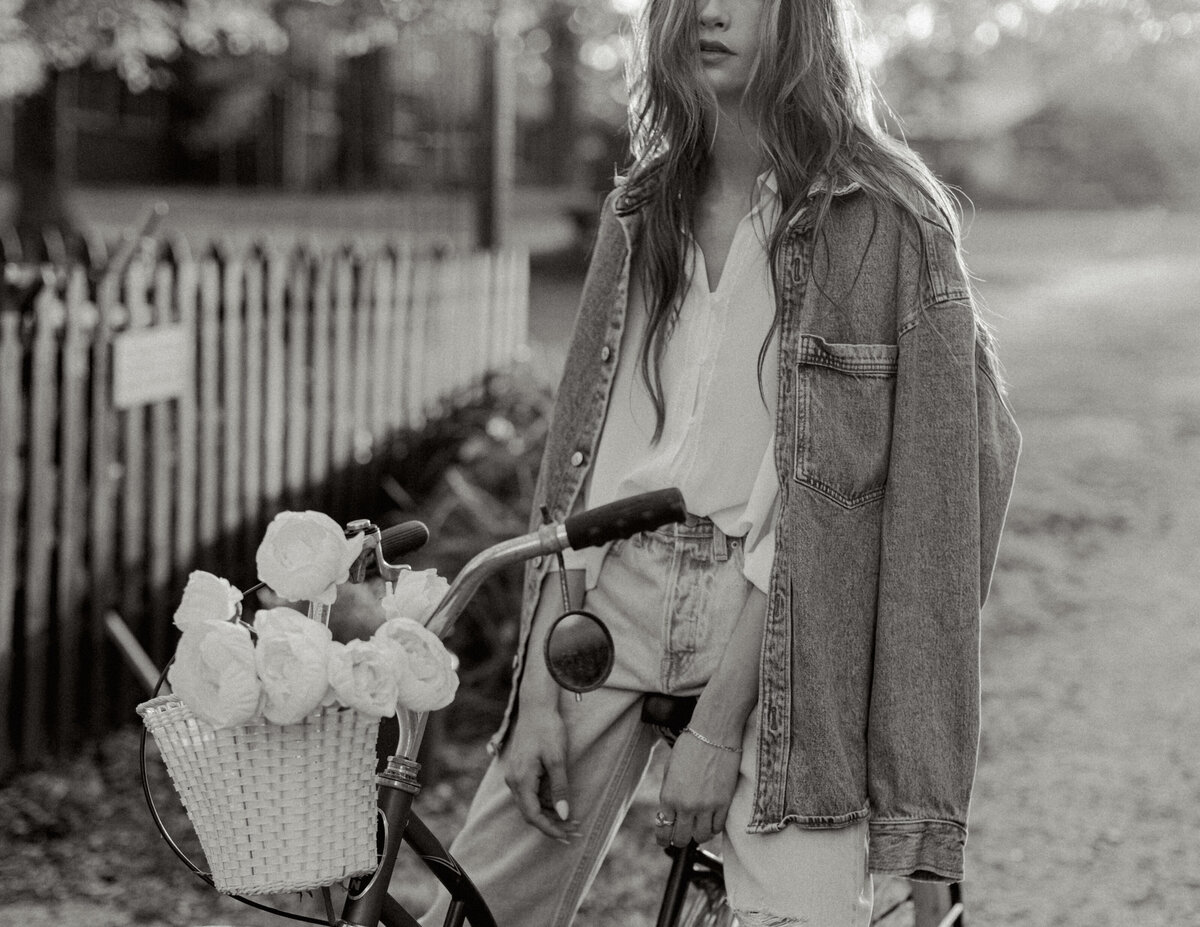 Woman in oversized denim jacket holding a vintage bicycle with white flowers in the basket at a countryside farm. Greenville SC branding photograph by Dani Lacey Photographs.