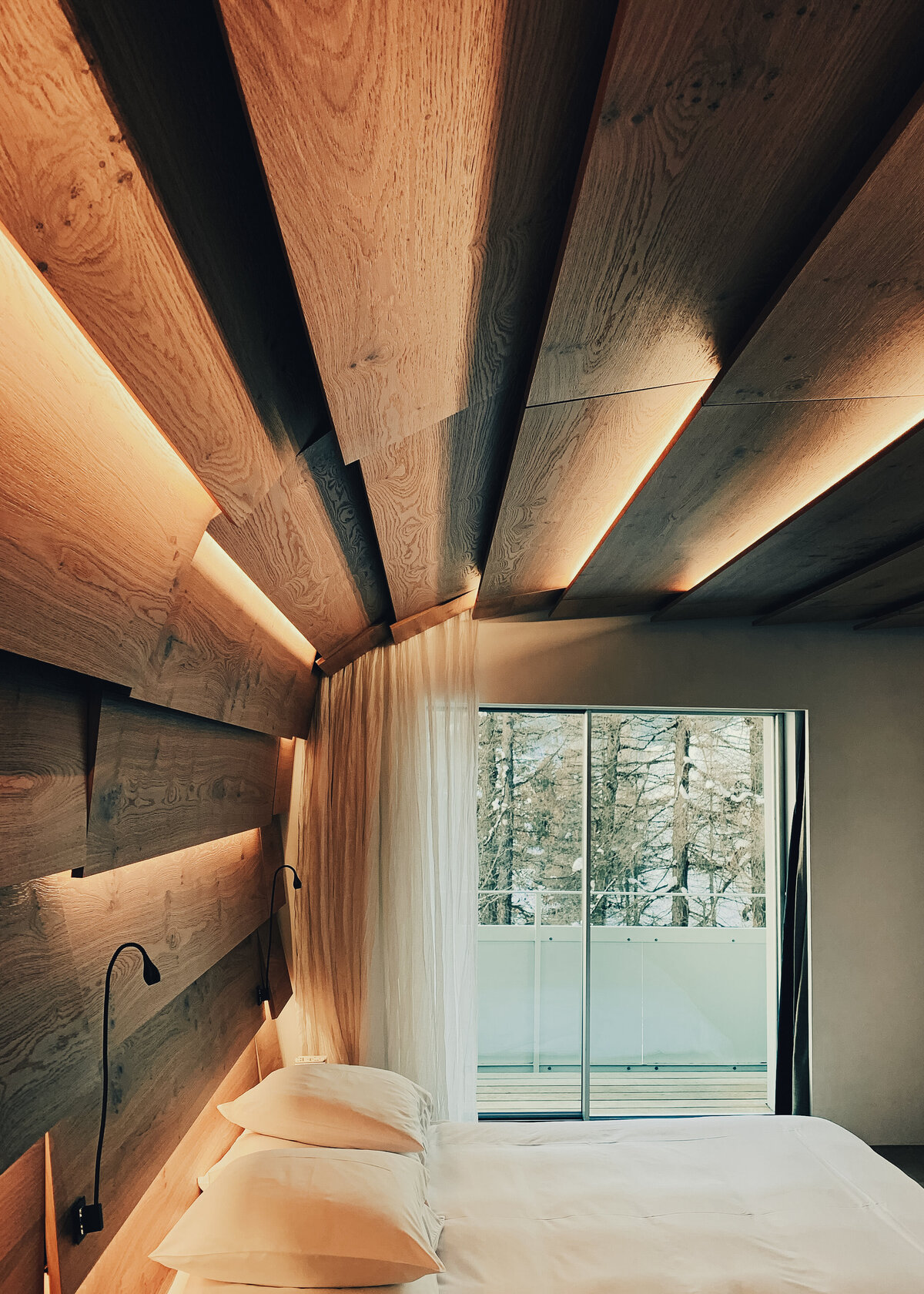 A ceiling is clad with natural oak with warm golden lighting in between with a white bed underneath.