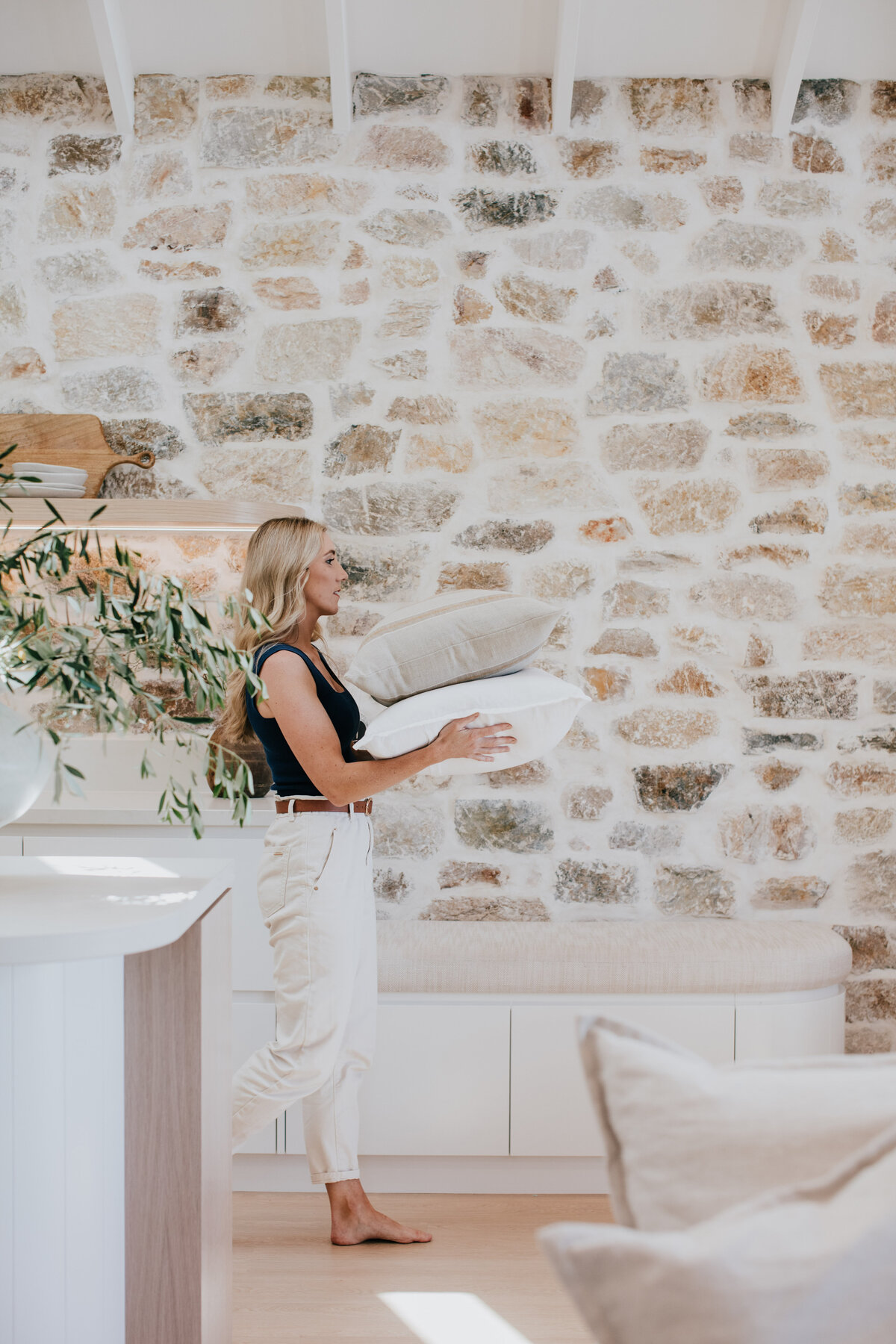 A person carrying pillows, smiling warmly in a bright room with a stone wall and wooden accents, echoes the touch of a Newcastle interior designer, known for their bespoke interior design flair that's truly unique to Newcastle NSW.