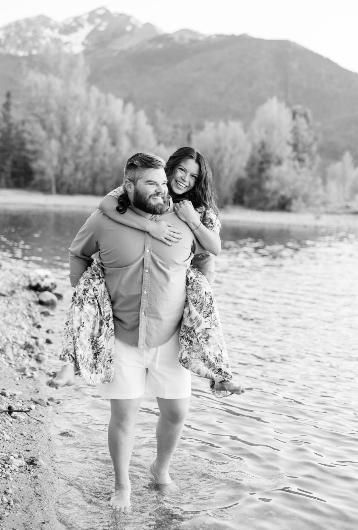 This is a black and white shot of the couple back down by the lake. The woman is on her boyfriends back, piggyback style, with her arms around his neck. They are smiling with one another. 