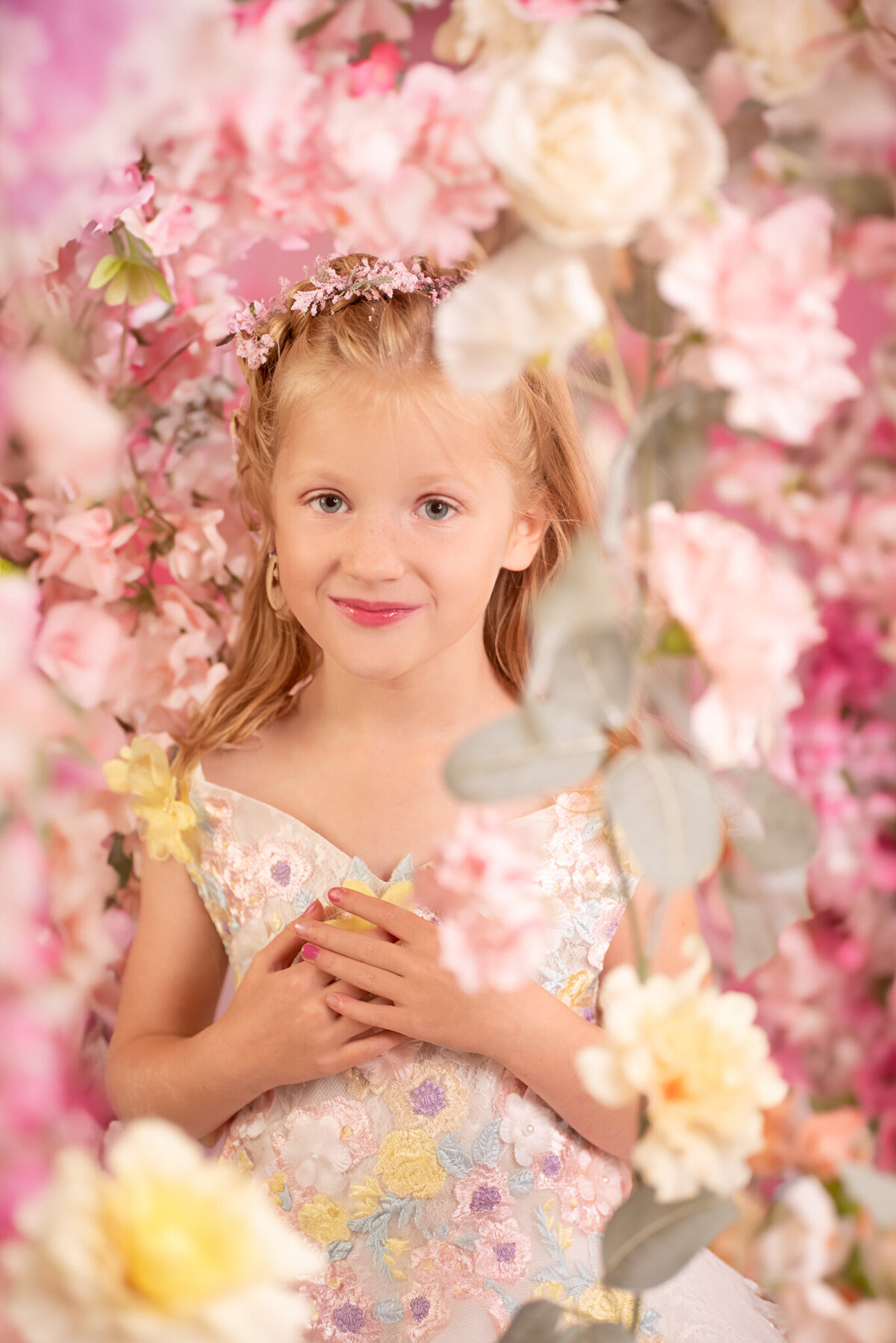 Whimsical portrait of a little girl dressed as a fairy with wings and surrounded by pink flowers. Captured in Overland Park, Kansas, this magical setup is ideal for creative child photography in the Kansas City area