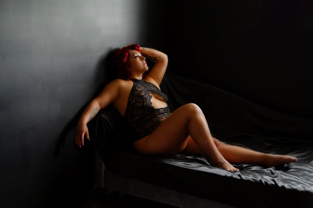 red haired woman sitting on a bed with black silk sheets with her hand in her hair for her Toronto boudoir photos