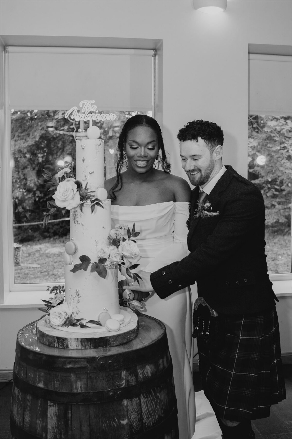 A bride and groom are cut their three tier wedding cake together in a bright space. The cake sits on an old barrel and guests take photos.