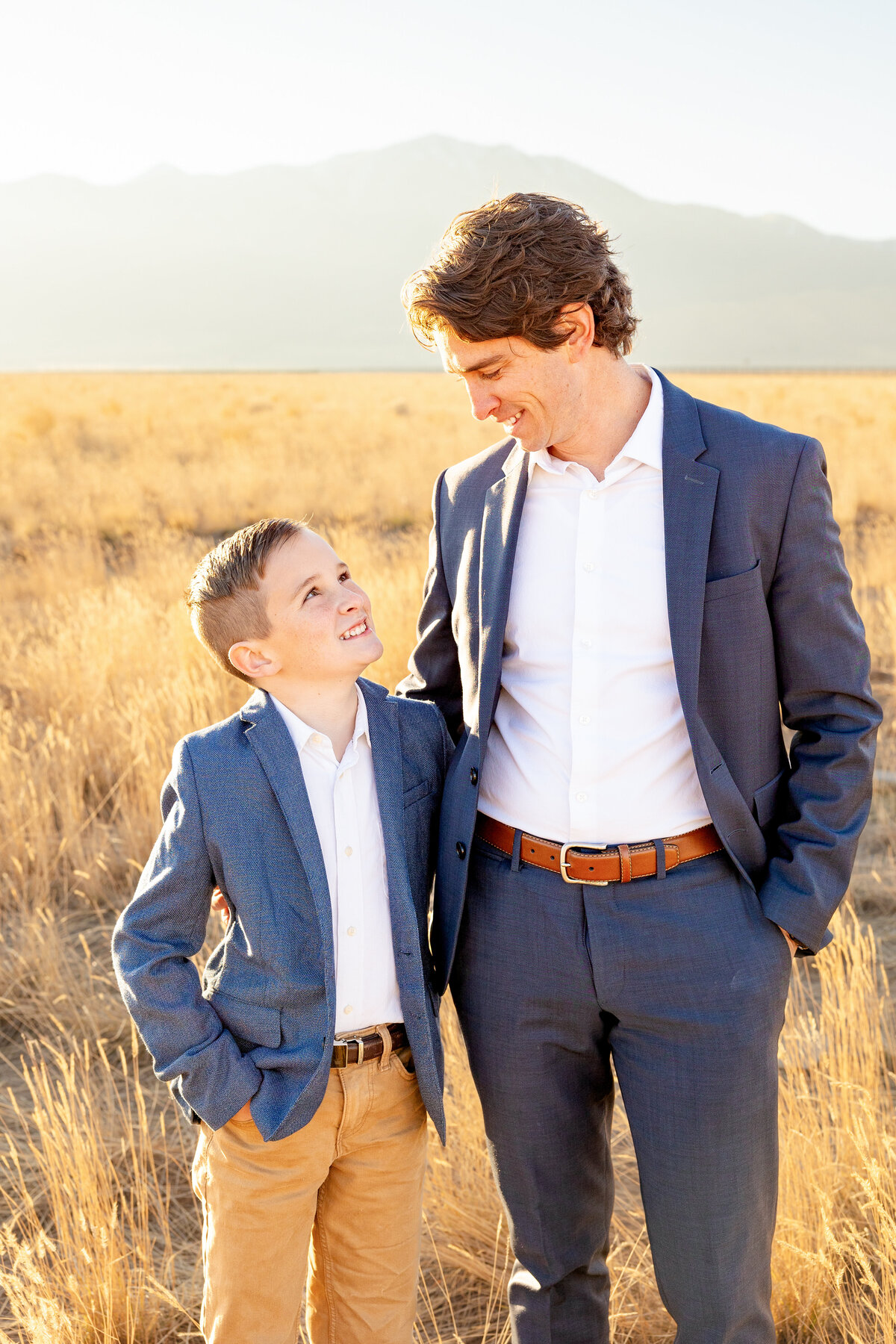 A father and son dressed in matching blue suits share a heartfelt gaze in a golden field with mountains in the background.