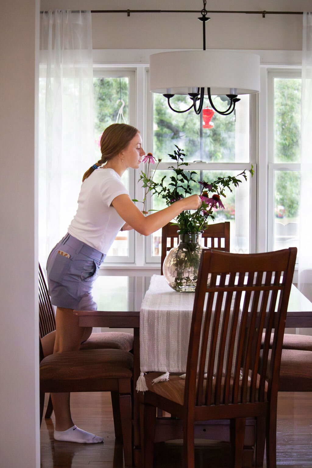 A woman with her hair pulled back in a ponytail, wearing a white t-shirt and light purple shorts, leans over a dark wooden dining table to arrange purple coneflowers and green leaves in a clear glass vase. The table is covered with a white runner and surrounded by wooden chairs with striped cushions. Large windows with sheer white curtains behind the table reveal a view of lush greenery. A black chandelier with a white drum shade hangs above the table.