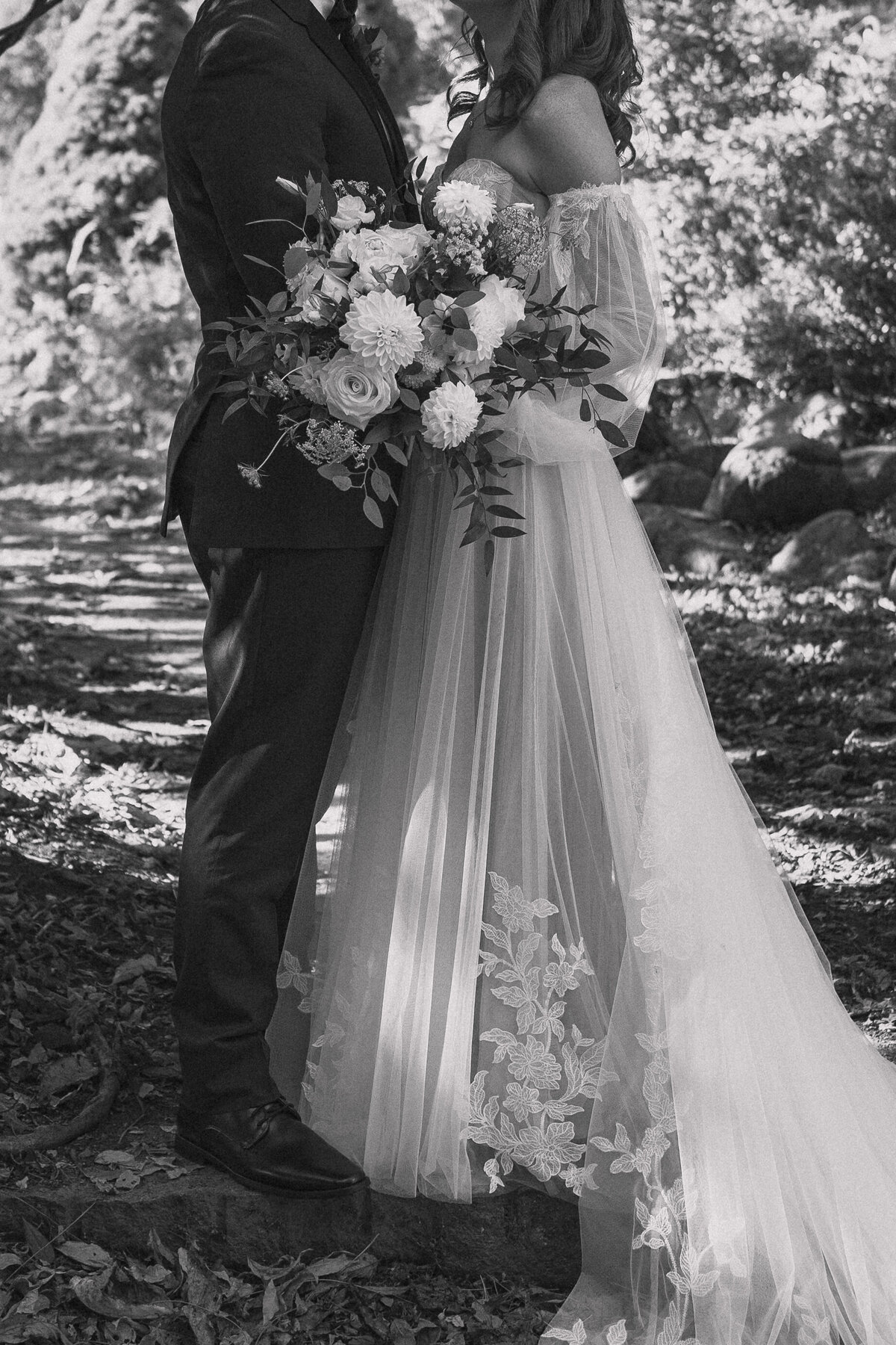 Black and white close-up photo of a bride and groom. The bride, in a flowing lace wedding dress, holds a large bouquet of flowers. The groom, dressed in a dark suit, stands close, embracing the bride. The image captures the intricate details of the bridal gown and the lush bouquet, set against a natural, outdoor background.