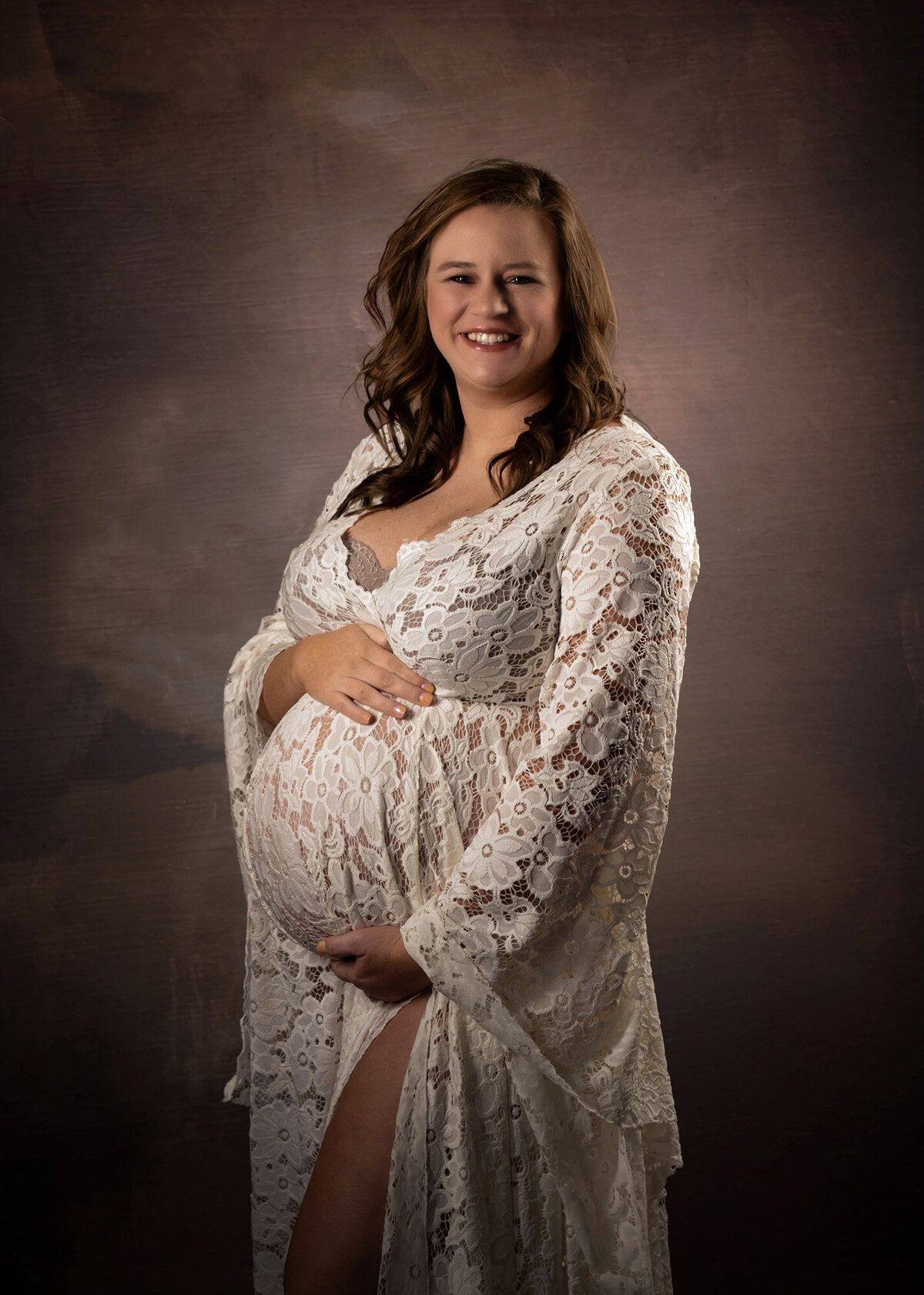 A mother to be stands in a studio in a white lace dress smiling with hands on her bump