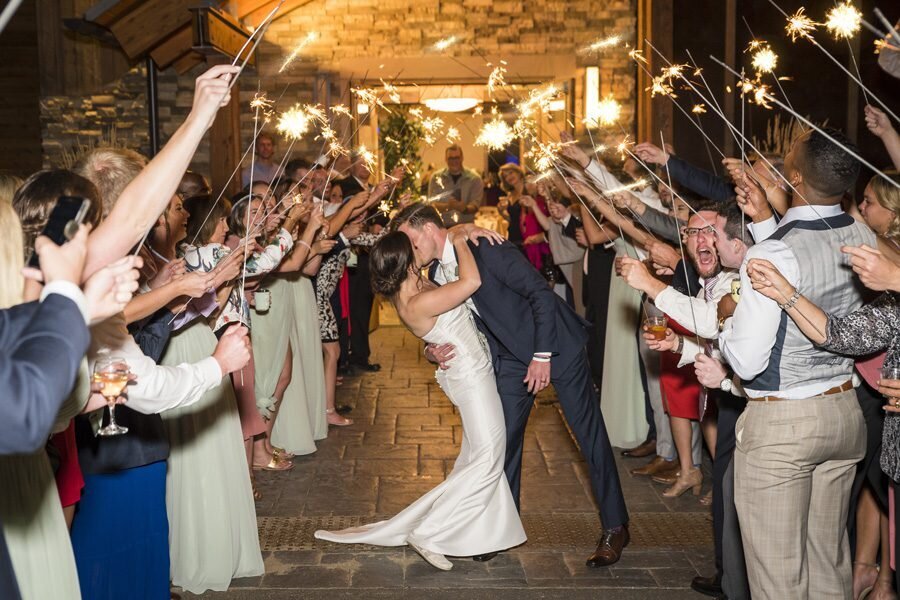 A groom dips his bride for a kiss  as they walk through their wedding guests who are holding sparklers.