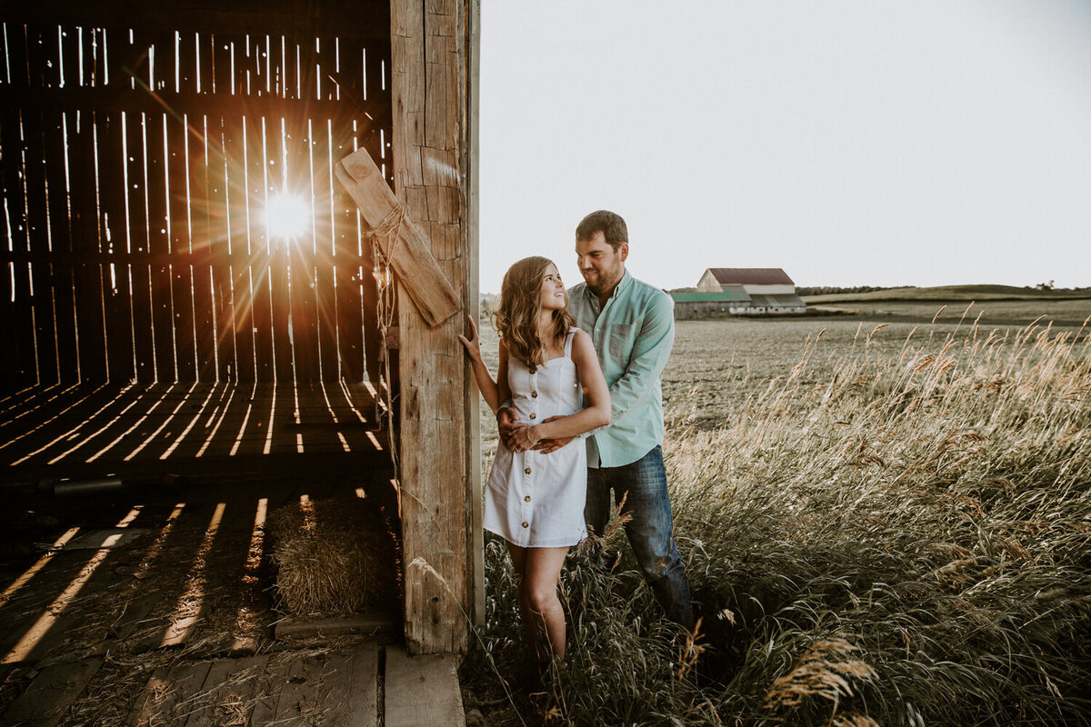 Woman and man standing in front of rustic barn for engagement session. The man is standing behind the woman with his arms around her waist. She is looking back at him. The sunset is peeking through the gaps in the barn boards.