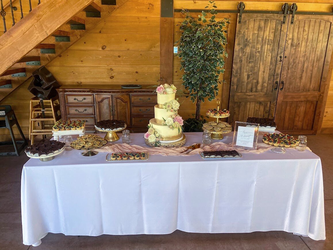 A large table holds a three tier wedding cake with painted gold bands around each tier and clusters of fresh flowers in pink and white. On either side of the cake are stands filled with mini pastries like fruit tarts and brownies, and mini cookies.