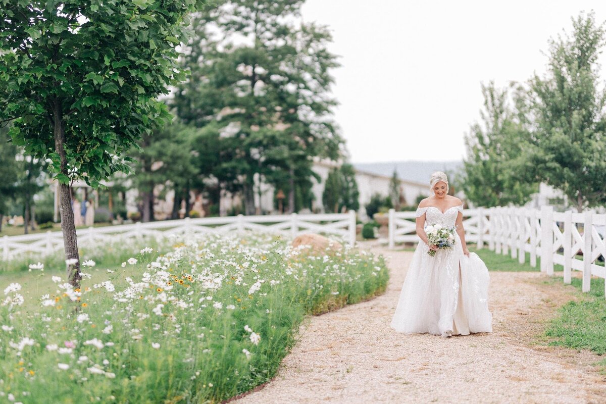 A bride holding a bouquet walking along a gravel path with white fences and blooming wildflowers.